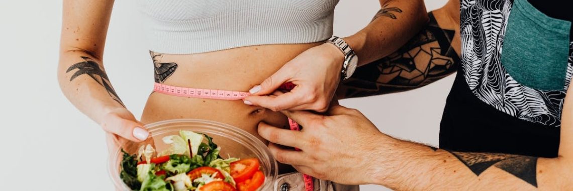 A couple measuring waistline with a tape and holding a fresh salad bowl, focusing on fitness and weight loss.